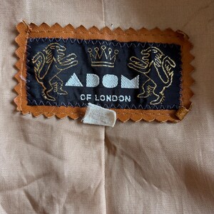 May include: Close-up of a label from a garment, featuring the text "ADOM OF LONDON". The label is black with gold detailing, including two lion figures and a crown. The label is framed by a brown, scalloped edge, set against a tan fabric background.