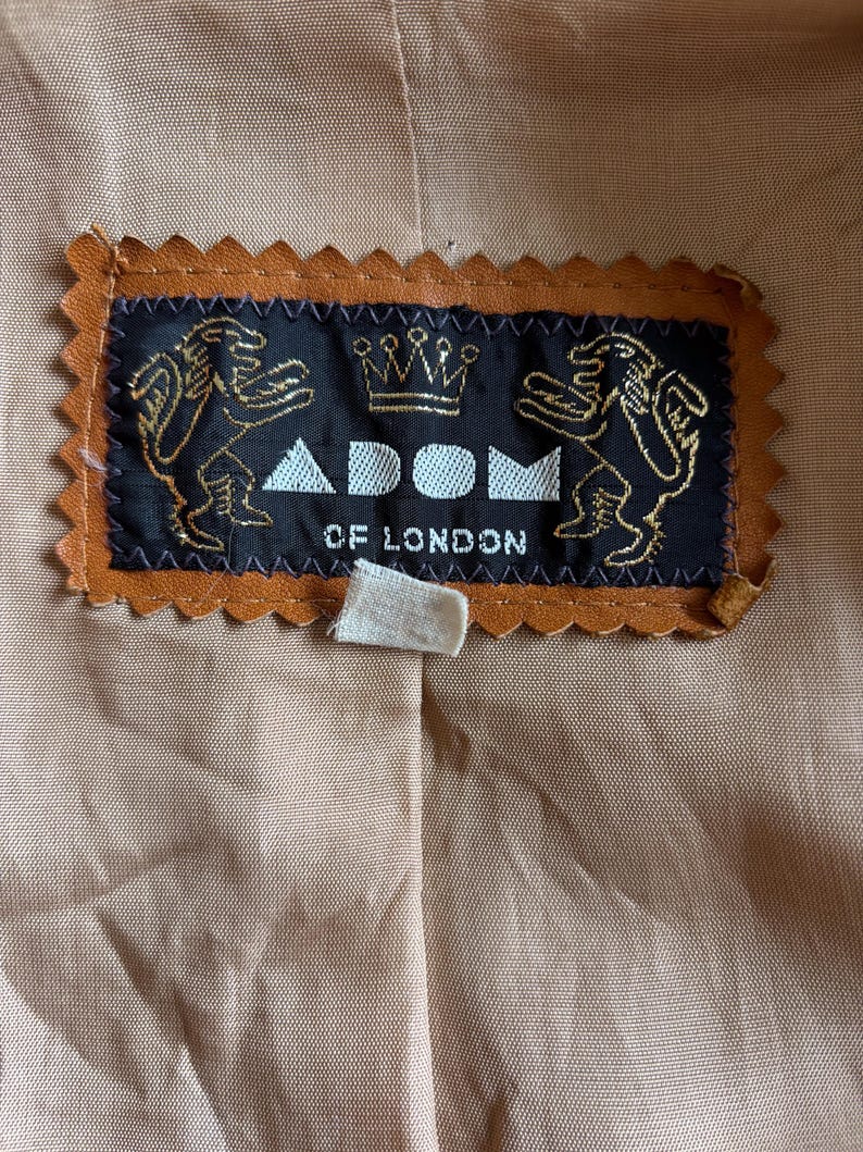 May include: Close-up of a label from a garment, featuring the text "ADOM OF LONDON". The label is black with gold detailing, including two lion figures and a crown. The label is framed by a brown, scalloped edge, set against a tan fabric background.
