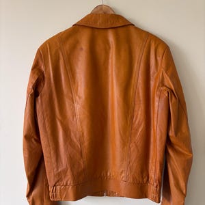 May include: A vintage brown leather jacket with a collared neckline and a zipper closure. The jacket is hanging on a wooden hanger against a neutral wall. The jacket's back features vertical stitching details.