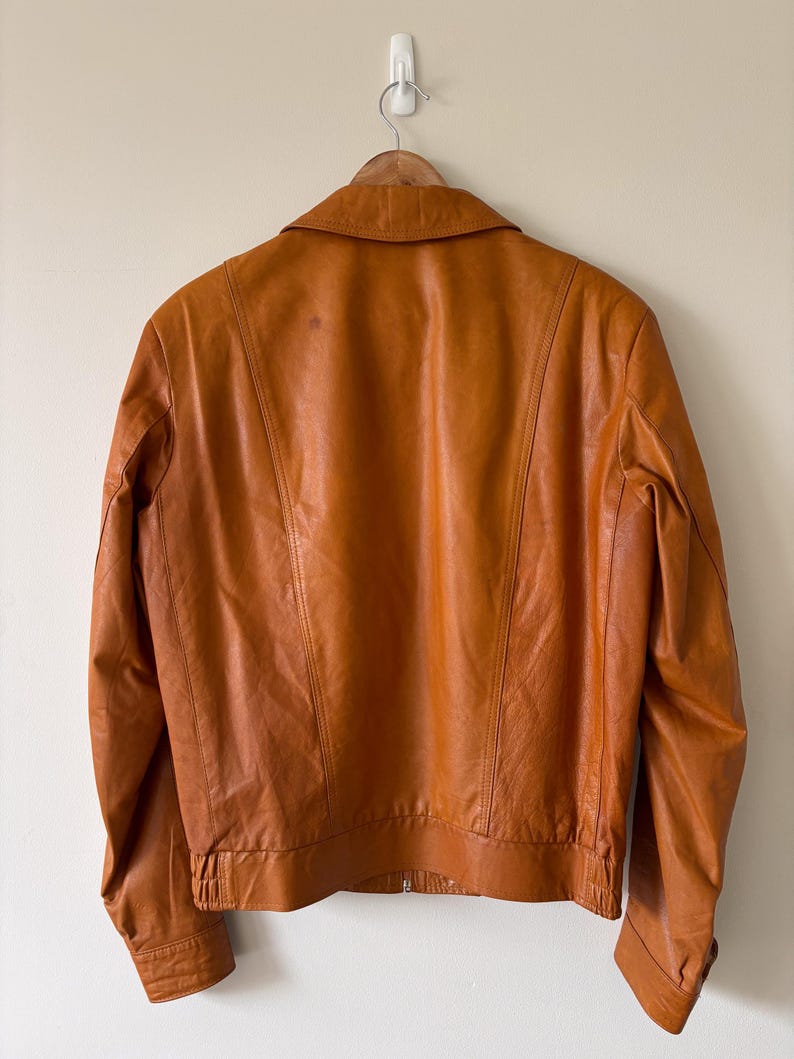 May include: A vintage brown leather jacket with a collared neckline and a zipper closure. The jacket is hanging on a wooden hanger against a neutral wall. The jacket's back features vertical stitching details.