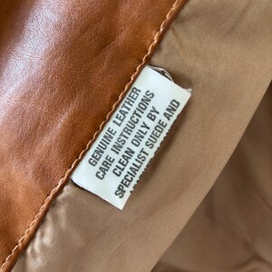 May include: Close-up of a brown leather garment with a tan lining. A white tag is visible, with the text "GENUINE LEATHER CARE INSTRUCTIONS CLEAN ONLY BY SPECIALIST SUEDE AND". The leather has a smooth texture.