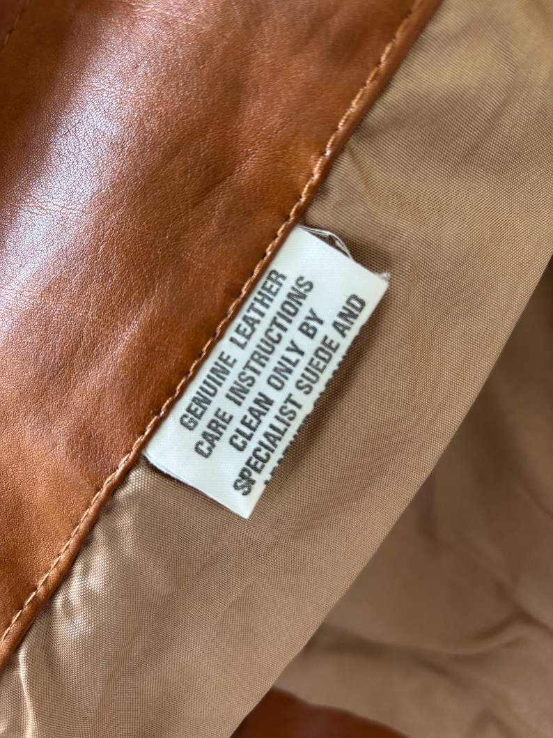 May include: Close-up of a brown leather garment with a tan lining. A white tag is visible, with the text "GENUINE LEATHER CARE INSTRUCTIONS CLEAN ONLY BY SPECIALIST SUEDE AND". The leather has a smooth texture.
