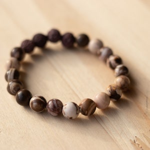 May include: A brown and white beaded bracelet with a dark brown lava rock bead. The bracelet is on a light brown wooden surface.
