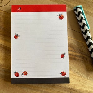 May include: A notepad featuring ladybug illustrations and a red border. The lined pages have ladybug designs in each corner and a black border at the bottom. A black and white chevron patterned pen rests on the side.