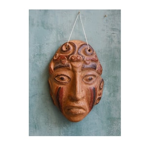 Puede incluir: Una máscara de terracota con intrincados detalles pintados, que presenta un rostro humano con una expresión seria. La máscara está colgada de una cuerda blanca.