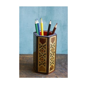 May include: A hexagonal wooden pencil holder with intricate inlaid geometric patterns in shades of brown, yellow, and orange. The holder is filled with coloured pencils, including red, blue, green, and yellow.