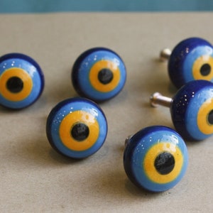 Op de afbeelding: Zes blauwe en gele keramische kastknoppen met een zwart oogontwerp. De knoppen hebben een zilveren metalen schroef aan de achterkant.