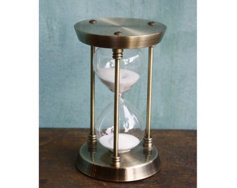 May include: A brass hourglass with white sand. The hourglass is on a wooden surface.