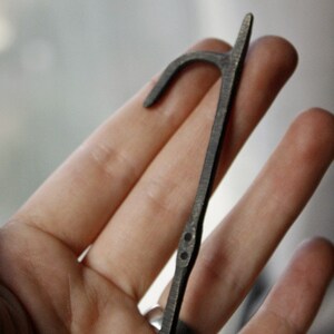 Puede incluir: Una herramienta de metal oscuro en forma de gancho con un extremo curvo y un mango recto. La herramienta tiene peque&ntilde;os detalles circulares y probablemente se utiliza para abrir o manipular algo. El dise&ntilde;o general es simple y funcional.