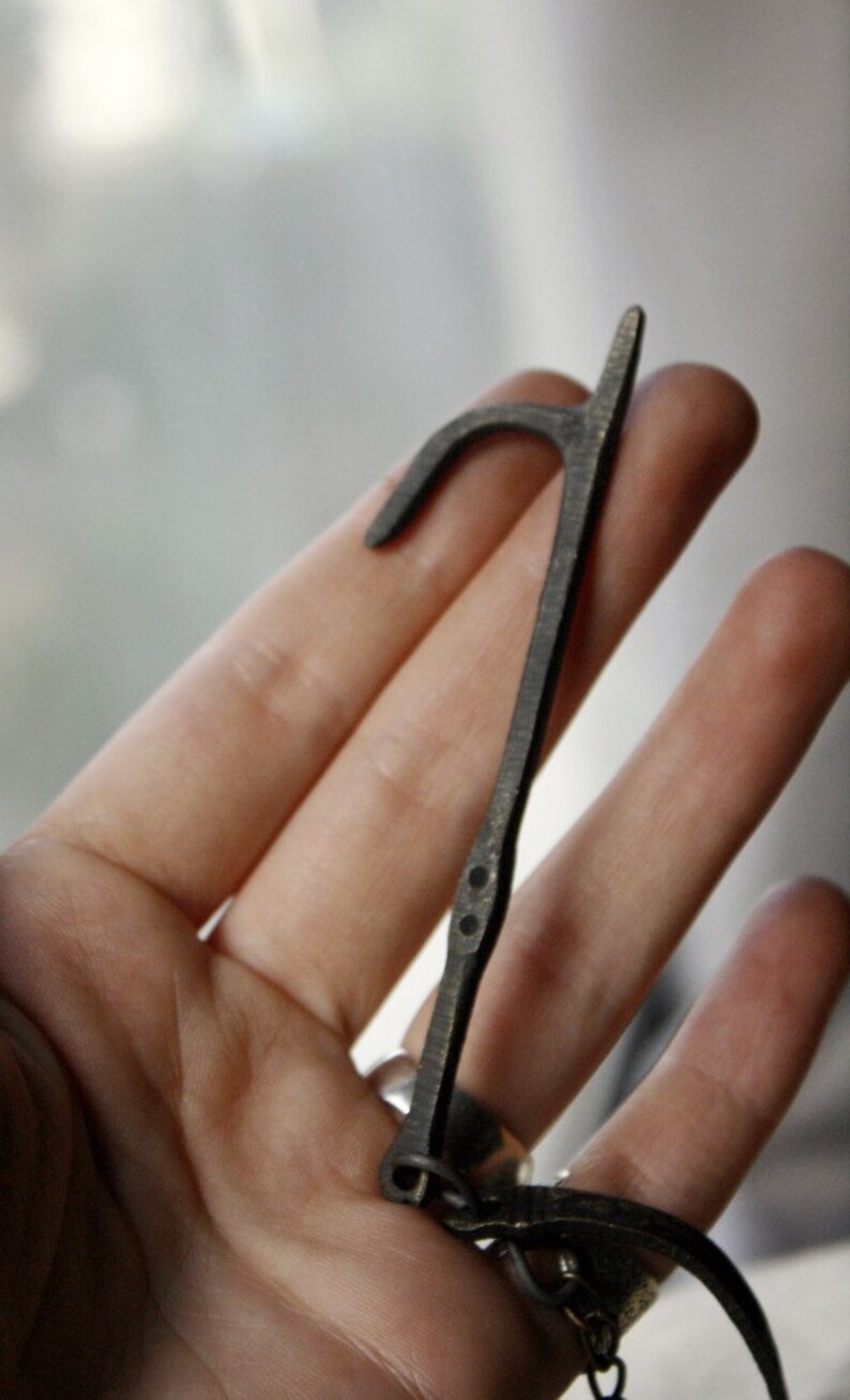 Puede incluir: Una herramienta de metal oscuro en forma de gancho con un extremo curvo y un mango recto. La herramienta tiene peque&ntilde;os detalles circulares y probablemente se utiliza para abrir o manipular algo. El dise&ntilde;o general es simple y funcional.
