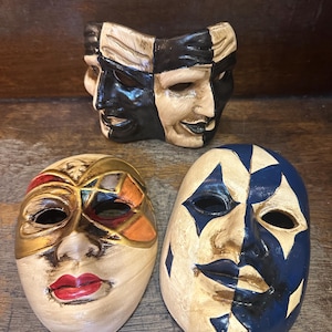 Peut inclure: Trois masques décoratifs aux designs distincts. Un masque présente une esthétique bicolore noir et crème. Un autre masque montre une base crème avec des motifs géométriques rouges, orange et dorés et des lèvres rouges. Le troisième a un motif en losanges bleu et crème.