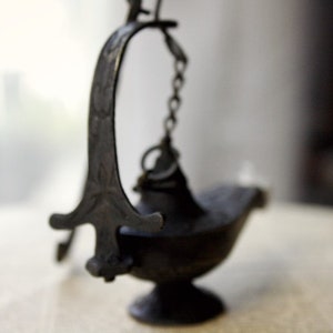 Puede incluir: Una l&aacute;mpara de aceite oscura de estilo antiguo con un asa curvada y una peque&ntilde;a cadena. La l&aacute;mpara tiene una base en forma de cuenco y un dise&ntilde;o decorativo y ornamentado. La l&aacute;mpara est&aacute; hecha de metal oscuro y se apoya sobre una superficie clara.