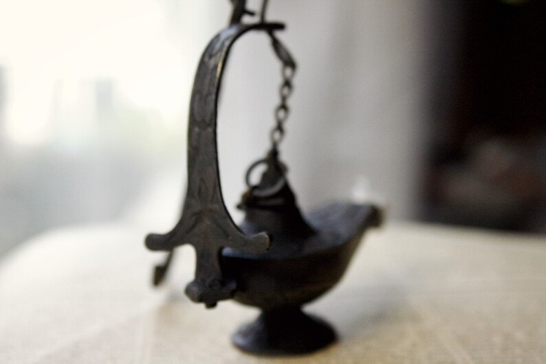 Puede incluir: Una l&aacute;mpara de aceite oscura de estilo antiguo con un asa curvada y una peque&ntilde;a cadena. La l&aacute;mpara tiene una base en forma de cuenco y un dise&ntilde;o decorativo y ornamentado. La l&aacute;mpara est&aacute; hecha de metal oscuro y se apoya sobre una superficie clara.
