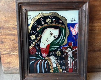Puede incluir: Una pintura enmarcada de un icono religioso que representa a la Virgen María con una túnica negra y verde, con un halo dorado, mirando hacia abajo a una pequeña representación de Jesús en la cruz. La pintura está sobre un fondo blanco con acentos azules y rosas.