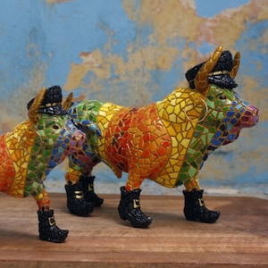 Puede incluir: Dos figuritas de toros de mosaico coloridos, una más grande que la otra, ambas con sombreros y botas negras. El toro más grande tiene un aro de oro en la oreja. Los toros están hechos de pequeñas baldosas de colores.