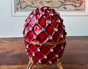 May include: A red jeweled egg-shaped trinket box with a gold trim and three gold legs. The box is decorated with a diamond pattern and rhinestones.