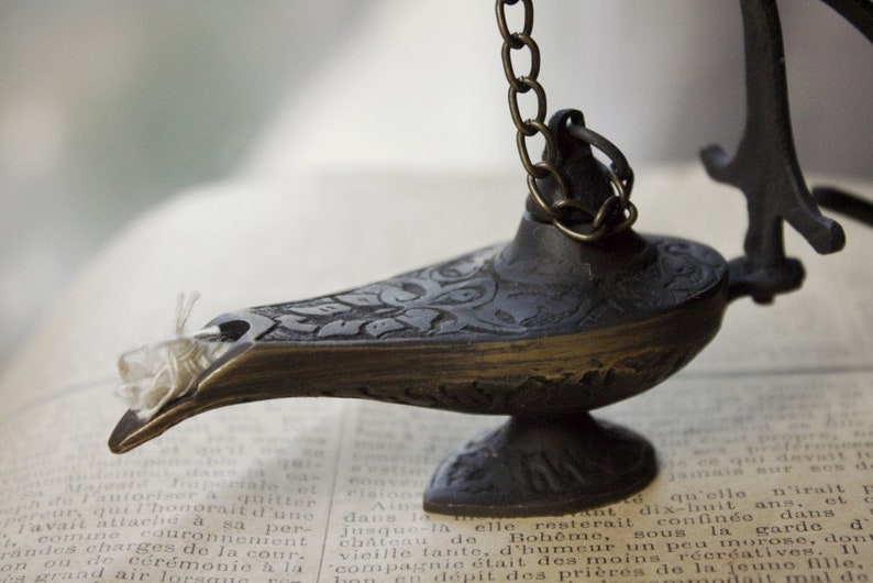 Puede incluir: L&aacute;mpara de genio antigua de color bronce con intrincados dise&ntilde;os florales. La l&aacute;mpara est&aacute; suspendida por una cadena y tiene un pico largo. Descansa sobre un libro antiguo con texto descolorido.