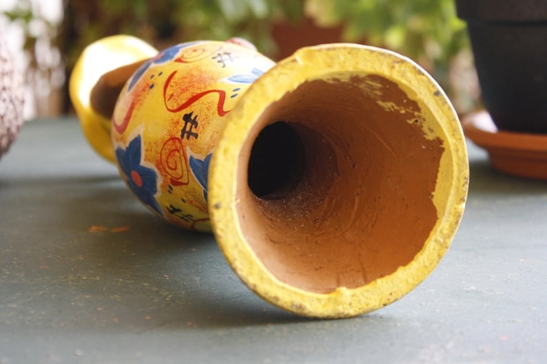 May include: A yellow ceramic vase with a painted design of red, blue, and orange flowers. The vase has a wide opening and a brown interior.