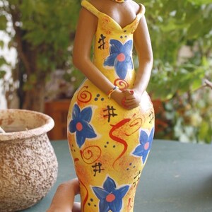 May include: A ceramic doll wearing a yellow hat and a yellow dress with blue and red floral patterns. The doll has brown skin and black hair.