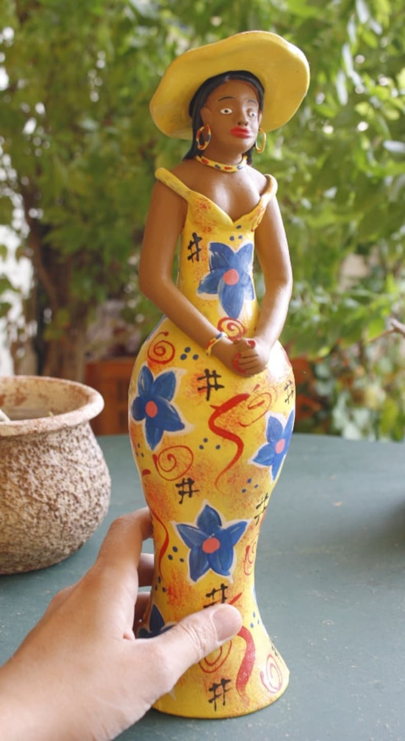 May include: A ceramic doll wearing a yellow hat and a yellow dress with blue and red floral patterns. The doll has brown skin and black hair.