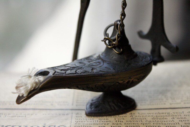 Puede incluir: Una l&aacute;mpara de aceite antigua y ornamentada, de color bronce oscuro. La l&aacute;mpara presenta intrincados dise&ntilde;os en relieve y una peque&ntilde;a cadena. Una mecha blanca sobresale de la boquilla. La l&aacute;mpara est&aacute; sobre un fondo de peri&oacute;dico antiguo.