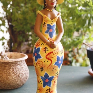 May include: A ceramic figurine of a woman wearing a yellow dress with blue and red floral patterns and a yellow hat. The figurine is standing on a green surface.