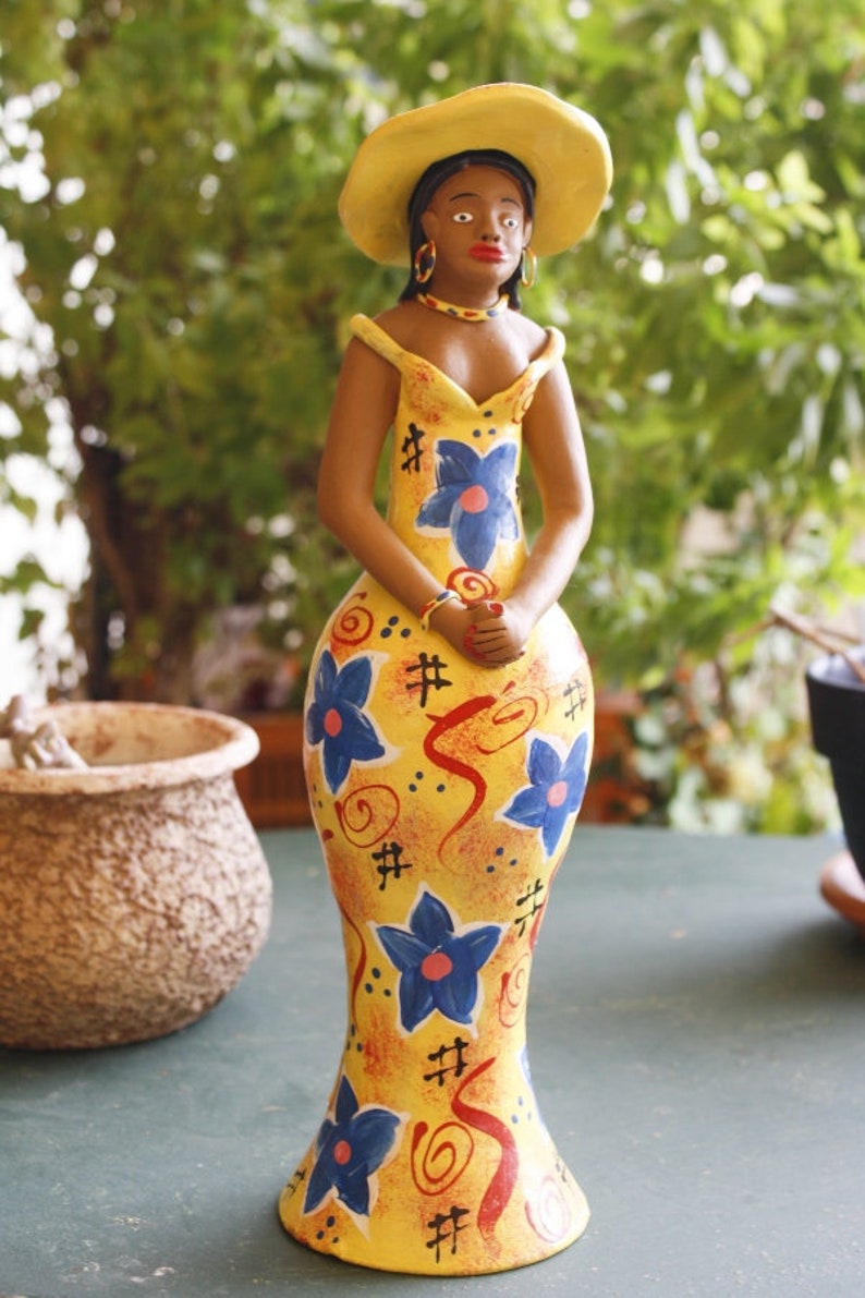 May include: A ceramic figurine of a woman wearing a yellow dress with blue and red floral patterns and a yellow hat. The figurine is standing on a green surface.