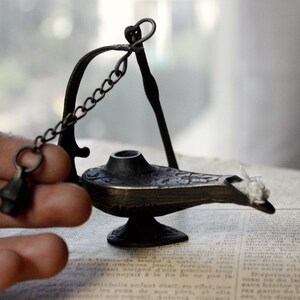 Puede incluir: Una peque&ntilde;a l&aacute;mpara de bronce de estilo antiguo con detalles intrincados. La l&aacute;mpara presenta un pico curvo, una peque&ntilde;a cadena y un asa decorativa. La l&aacute;mpara est&aacute; sobre un libro antiguo, lo que sugiere un tema de historia o fantas&iacute;a.