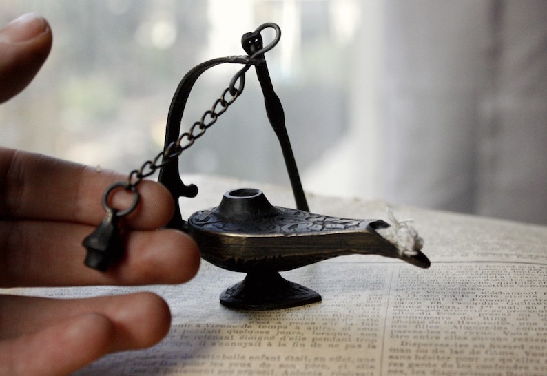 Puede incluir: Una peque&ntilde;a l&aacute;mpara de bronce de estilo antiguo con detalles intrincados. La l&aacute;mpara presenta un pico curvo, una peque&ntilde;a cadena y un asa decorativa. La l&aacute;mpara est&aacute; sobre un libro antiguo, lo que sugiere un tema de historia o fantas&iacute;a.