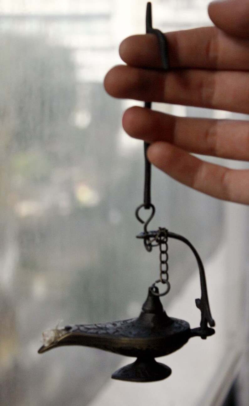 Puede incluir: Una peque&ntilde;a l&aacute;mpara de bronce de estilo antiguo con un pico curvo y un asa decorativa. La l&aacute;mpara est&aacute; suspendida por una cadena y un gancho. El dise&ntilde;o de la l&aacute;mpara recuerda a una l&aacute;mpara de genio.