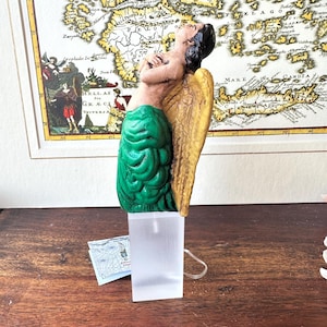 May include: A green and gold angel figurine with wings outstretched, looking up. The figurine is made of painted ceramic and is standing on a clear acrylic base.