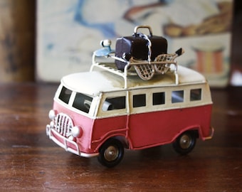 May include: A red and white toy van with a white roof and a luggage rack on top. The luggage rack is holding a brown suitcase, a blue and white beach umbrella, and a white and blue beach net. The van has a white front bumper and a black grill.