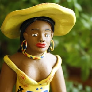 May include: A ceramic figurine of a woman wearing a yellow hat and a yellow dress with red and blue accents. The figurine has brown skin and is wearing large hoop earrings.