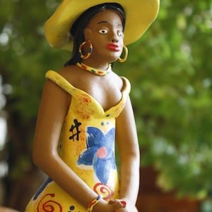May include: A ceramic figurine of a woman wearing a yellow hat and a yellow dress with blue and red floral designs. The figurine has brown skin and is wearing large gold earrings.