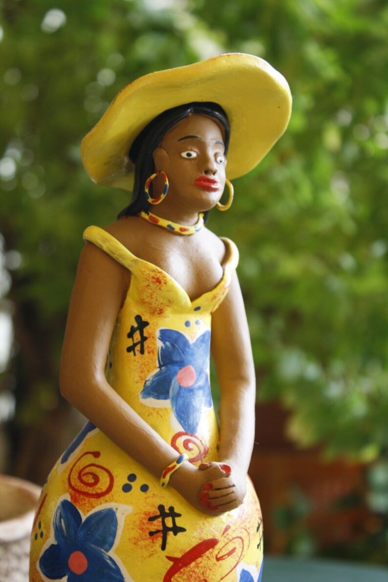 May include: A ceramic figurine of a woman wearing a yellow hat and a yellow dress with blue and red floral designs. The figurine has brown skin and is wearing large gold earrings.