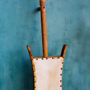 Puede incluir: Un instrumento musical de madera con una caja de resonancia de cuero blanco y un mango de madera tallado. El instrumento est&aacute; de pie sobre una superficie de madera.