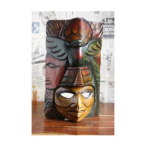 Op de afbeelding: Een kleurrijk houten masker met een gesneden gezicht en een vogel erop. Het masker is geschilderd in tinten rood, groen, geel en bruin.