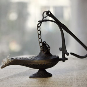 Puede incluir: Una l&aacute;mpara de genio de color bronce con una boquilla curva y detalles ornamentados. La l&aacute;mpara presenta una cadena y un asa con bisagras. La l&aacute;mpara est&aacute; sobre un libro con texto.