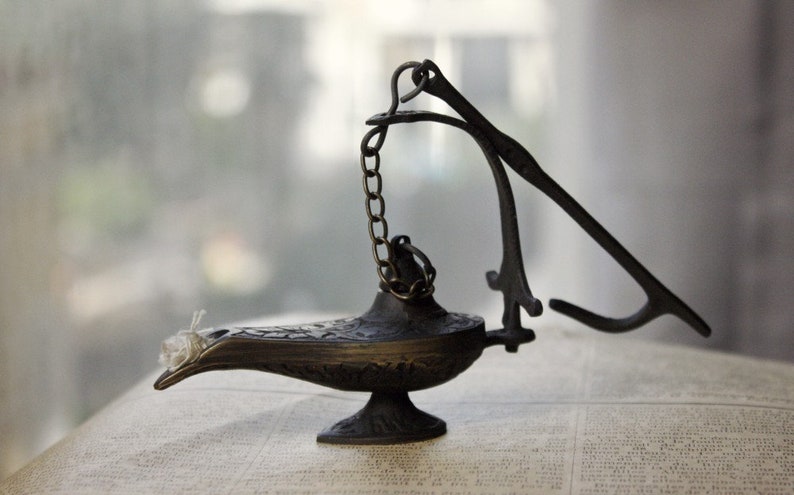Puede incluir: Una l&aacute;mpara de genio de color bronce con una boquilla curva y detalles ornamentados. La l&aacute;mpara presenta una cadena y un asa con bisagras. La l&aacute;mpara est&aacute; sobre un libro con texto.
