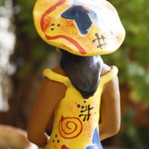 May include: A ceramic figurine of a person wearing a yellow, red, and blue hat with a colorful, abstract design. The figurine is wearing a yellow dress with red, blue, and orange painted details.