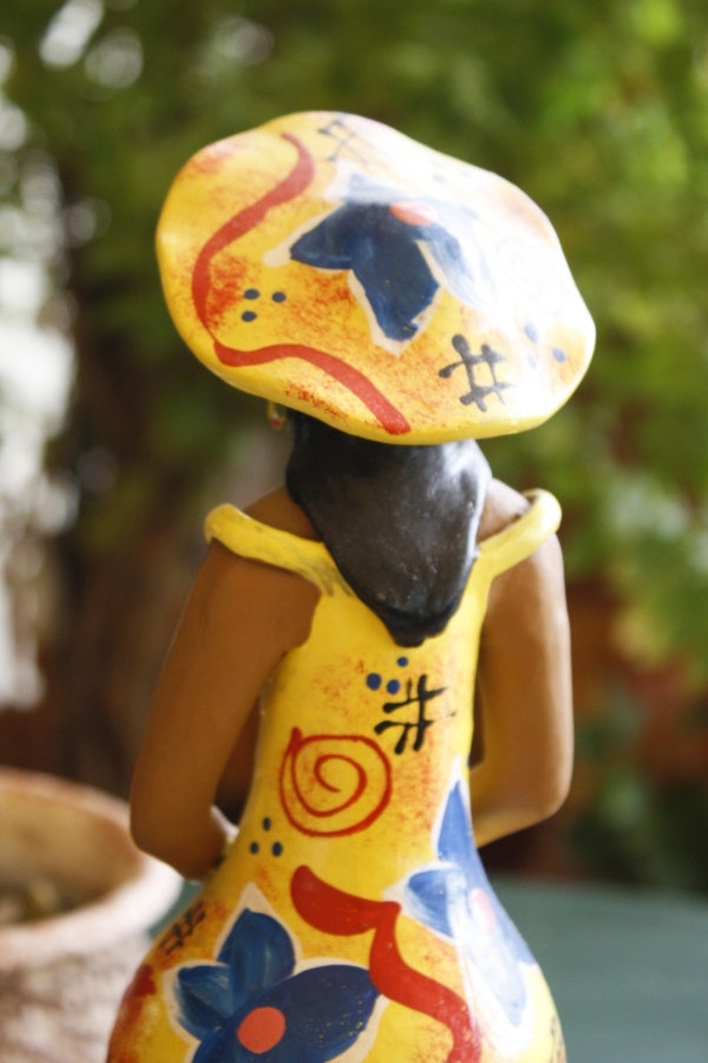 May include: A ceramic figurine of a person wearing a yellow, red, and blue hat with a colorful, abstract design. The figurine is wearing a yellow dress with red, blue, and orange painted details.