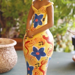 May include: A ceramic figurine of a woman wearing a yellow hat and a yellow dress with blue and red floral patterns. The figurine is painted in a whimsical style.