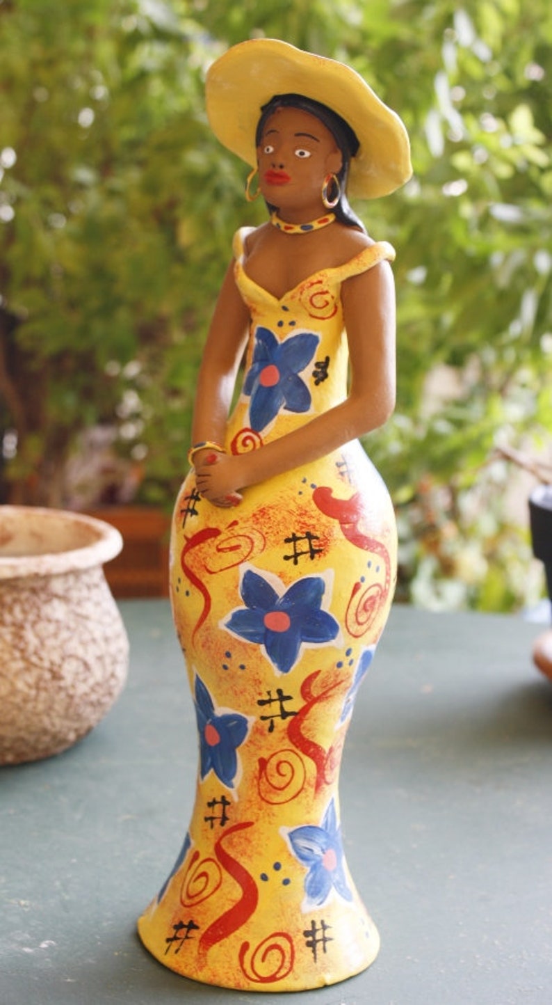 May include: A ceramic figurine of a woman wearing a yellow hat and a yellow dress with blue and red floral patterns. The figurine is painted in a whimsical style.
