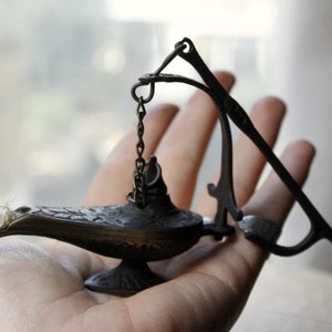 Puede incluir: Una peque&ntilde;a l&aacute;mpara de estilo antiguo con un pico curvo y detalles ornamentados. La l&aacute;mpara es de color bronce oscuro y tiene una cadena unida a un asa curva. Una mecha blanca es visible en el pico. La l&aacute;mpara se sostiene en una mano.