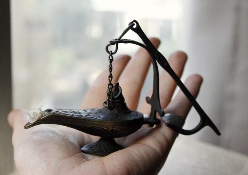 Puede incluir: Una peque&ntilde;a l&aacute;mpara de estilo antiguo con un pico curvo y detalles ornamentados. La l&aacute;mpara es de color bronce oscuro y tiene una cadena unida a un asa curva. Una mecha blanca es visible en el pico. La l&aacute;mpara se sostiene en una mano.