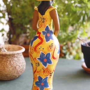 May include: A yellow ceramic figurine of a woman wearing a yellow and blue patterned dress and a yellow hat with a blue and red design. The figurine has a stylized, abstract design.