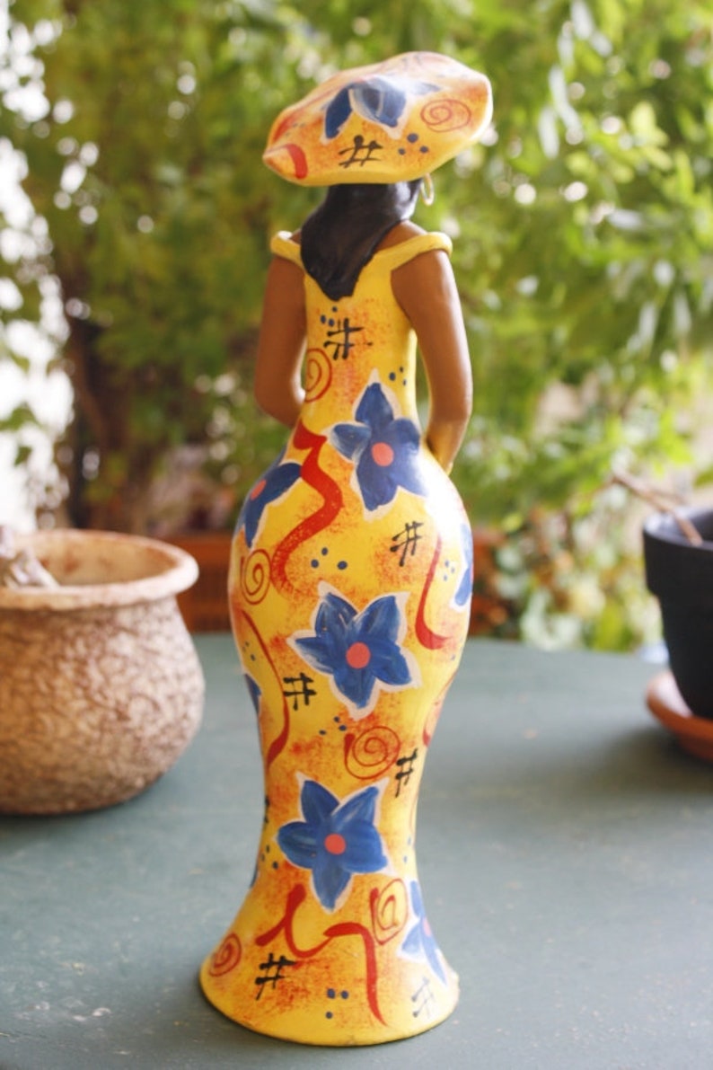 May include: A yellow ceramic figurine of a woman wearing a yellow and blue patterned dress and a yellow hat with a blue and red design. The figurine has a stylized, abstract design.