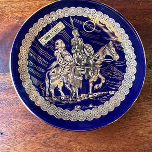 Może przedstawiać: Błękitny talerz ceramiczny z ozdobną obwódką. Na talerzu przedstawiono scenę z dwoma osobami na koniach z napisem "ORO Y LEY" na banerze.