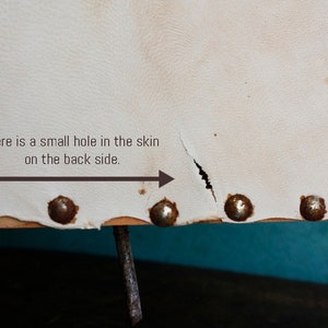 Puede incluir: Silla de cuero blanco con un peque&ntilde;o agujero en la parte posterior. La silla tiene un marco de madera y tachuelas met&aacute;licas alrededor de los bordes.