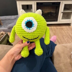 May include: A green crocheted plush toy with one large blue eye and a white circle around it. The toy has two small horns on top of its head and two small arms and legs.